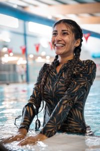 Summaya is standing at the edge of the swimming pool with her hands placed on the side of the pool, wearing her hair in two braids and smiling.