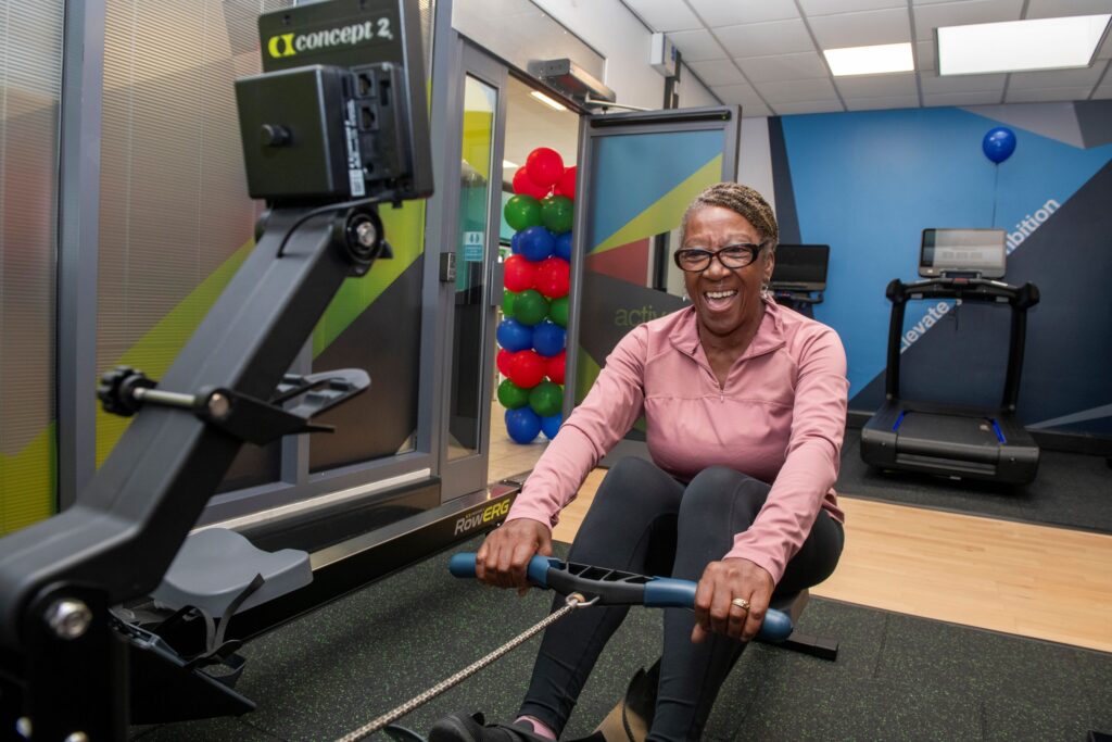 Cllr Jenkins, in a blush coloured workout top, is sat on a self-powered rowing machine, in the Self Powered workout zone. 