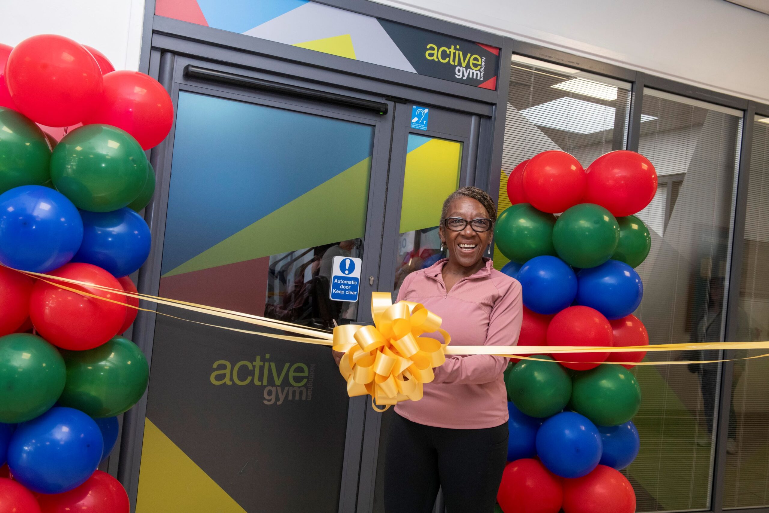 Cllr Jenkins stands behind a gold ribbon with the entrance to the newly refurbished gym behind. Red, Green and blue balloons are decorated to both side of the door.