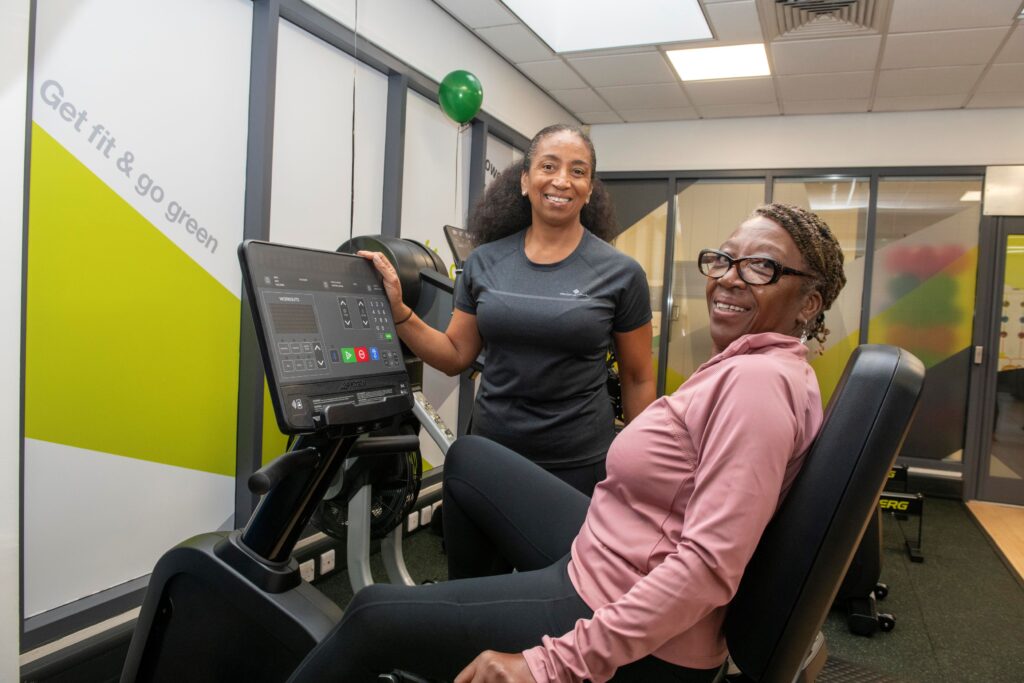 Cllr Jenkins, in a blush coloured workout top, is sat on an upright bike, in the Self Powered workout zone. 