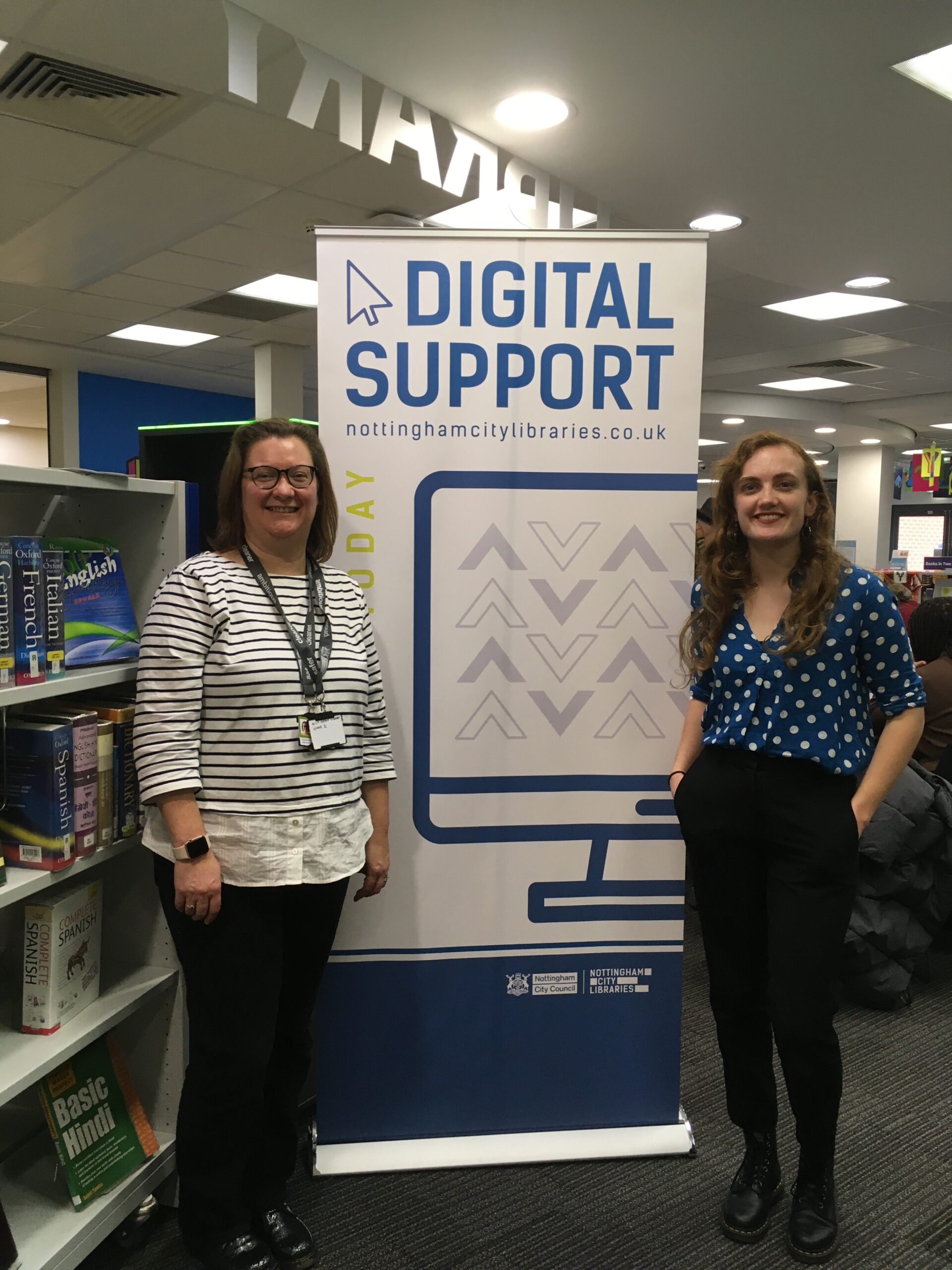 two female library staff members stook smiling next to a banner that says Digital Support Here Today