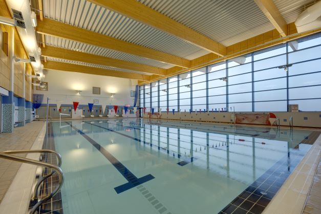 Indoor swimming pool with clear water, lane markings on the pool floor, colourful pennant flags above the lanes, large windows along one side, and a high wooden-beam ceiling letting in natural light.