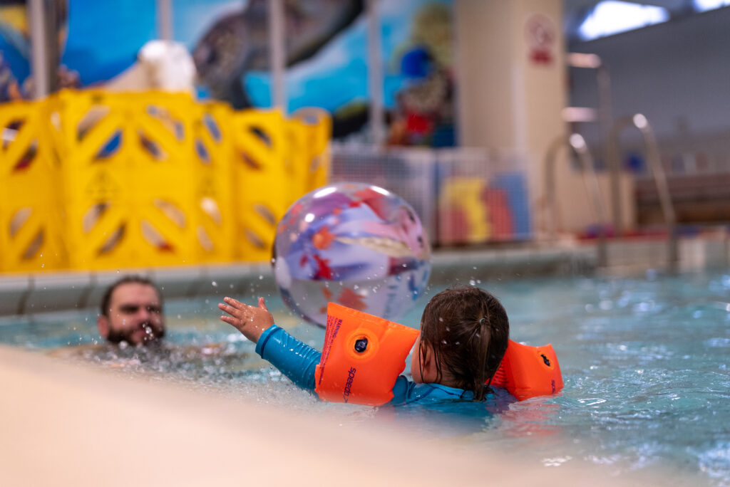 A young child in a leisure pool reaches toward a patterned inflatable ball, wearing orange armbands, with an adult nearby in the water